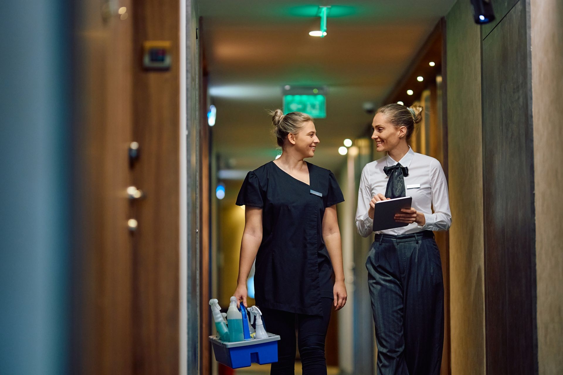 Two hotel staff walking in a corridor, one holding cleaning supplies, the other a tablet.