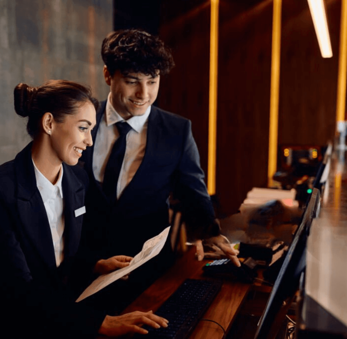 Two people in business suits working at a desk with a computer and documents in an office setting.